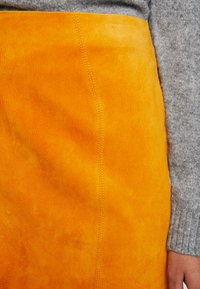 Gonna in finta suede arancione con una texture morbida, caratterizzata da una cucitura visibile lungo il lato. Abbinata a un maglione grigio in maglia.