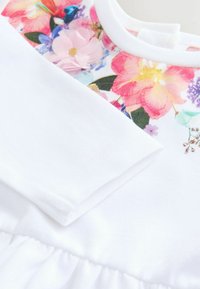 Hvid langærmet beklædningsgenstand med et blomsterprintpanel i toppen med rosa, lilla og blå blomster med grønne blade.