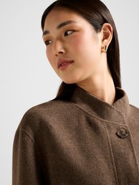 Rudos spalvos vilnos mišinio paltas su aukštu apykakliu, su vienu mygtuko užsegimu ir minkštu, tekstūruotu paviršiumi. Akcentuotas minimalistišku dizainu.