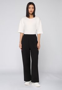 Witte oversized t-shirt met korte mouwen, gecombineerd met zwarte broek met wijde pijpen en gestikte details. Schoenen zijn wit met een textuurloze zool.