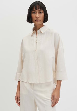 Vrouw met kort zwart haar, die een oversized crèmekleurig overhemd met knopen en bijpassende broek draagt tegen een effen witte achtergrond.