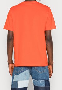 Mann in einem orangefarbenen kurzärmeligen T-Shirt und geflickten Jeansshorts, der mit dem Rücken zur Kamera vor einem einfarbigen Hintergrund steht.