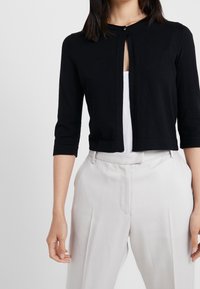 Schwarze, kurz geschnittene Strickjacke mit rundem Ausschnitt und drei Viertelärmeln. Besitzt einen kleinen Frontknopf und eine glatte, weiche Strickstruktur.