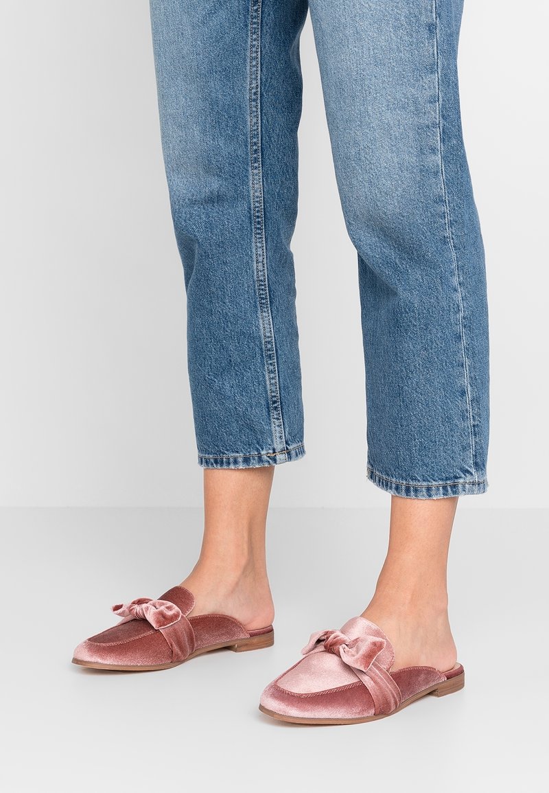 Pieds portant des mocassins en velours rose à enfiler avec un détail de nœud, associés à un jean denim bleu raccourci sur un fond clair uni.