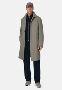 Cappotto lungo grigio chiaro con bottoni sul davanti, due tasche laterali, indossato sopra un maglione con zip blu navy. Il modello indossa anche pantaloni blu navy e scarpe da ginnastica grigie.