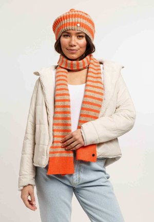 Gehaakte oranje en beige gestreepte muts met een pompoen, gecombineerd met een bijpassende gestreepte sjaal. Het model draagt een crèmekleurige pufferjack en lichte jeans.