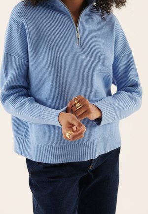 Person iført en lyseblå ribstrikket sweater med halv lynlås og mørke bukser, med flere guldringe på fingrene.