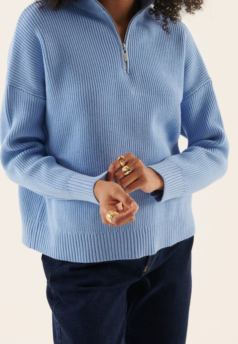Personne portant un pull côtelé bleu clair avec une demi-fermeture éclair et un pantalon foncé, avec plusieurs bagues en or aux doigts.