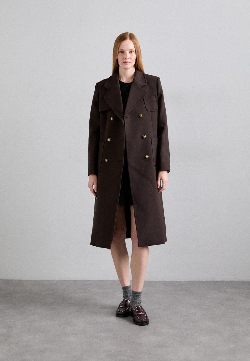 Manteau marron croisé en laine avec de grands revers et des boutons dorés ; associé à une robe noire et des chaussures sombres à motifs.