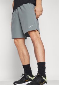 Graue Sportshorts mit einem Seitenschlitz, versehen mit einem dezenten Logo. Getragen mit schwarzen Socken und schwarzen Sportschuhen mit grünen Akzenten.