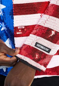 Rode en witte paillettenjas met strepen, voorzien van een blauwe paillettenpatch met witte sterren. Zwarte "SUITMEISTER" label op de mouw.