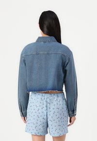 Κοντοκομμένο μπλε τζιν μπουφάν με φθαρμένο στρίφωμα, μακριά μανίκια και κουμπωτά μπατζάκια· συνδυασμένο με ανοιχτό μπλε σορτς με λουλούδια.