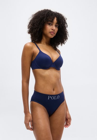 Γυναίκα με κατσαρά μαλλιά που φοράει σκούρο μπλε σουτιέν και αντίστοιχο ψηλόμεσο σορτς με το λογότυπο "POLO", στέκεται μπροστά σε απλό ανοιχτόχρωμο φόντο.