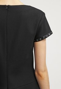 Musta mekko lyhyillä hihoilla, jossa on saumatön takakaukalo ja pyöreät rei'ityksensä hihojen reunoilla. Tasainen kangasrakennus.