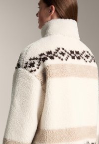 Biała pluszowa flanelowa kurtka z wysokim kołnierzem, ozdobiona brązowymi geometrycznymi wzorami i beżowymi paskami na środku. Miękka tekstura.