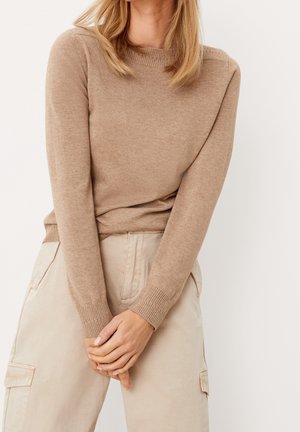 Femme aux cheveux blonds mi-longs portant un pull en tricot beige et un pantalon cargo de couleur claire, les mains jointes devant elle.