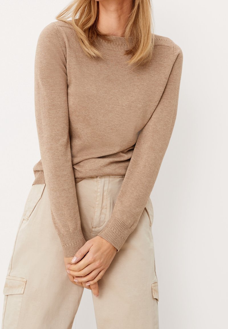 Femme aux cheveux blonds mi-longs portant un pull en tricot beige et un pantalon cargo de couleur claire, les mains jointes devant elle.