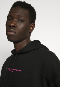 Black hoodie with a drawstring, featuring bold pink text on the front. Cotton blend fabric with a smooth texture.