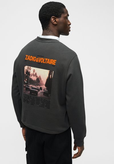 Άνδρας που φοράει σκουρόχρωμο γκρι φούτερ με μια σκηνή vintage αυτοκινήτου στο δρόμο και έντονο πορτοκαλί κείμενο "ZADIG & VOLTAIRE" στην πλάτη, κοιτάζοντας πλάγια.