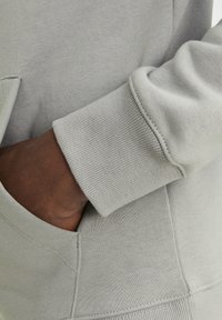 Light grey hoodie fabric with ribbed cuffs. Close-up shows textured surface and stitching details. Side pocket visible, providing functionality.