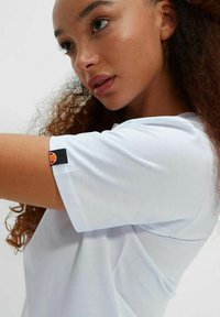 Camiseta de algodón blanca con mangas cortas, que presenta una etiqueta negra con un logo rojo en la manga izquierda, textura suave y corte relajado.