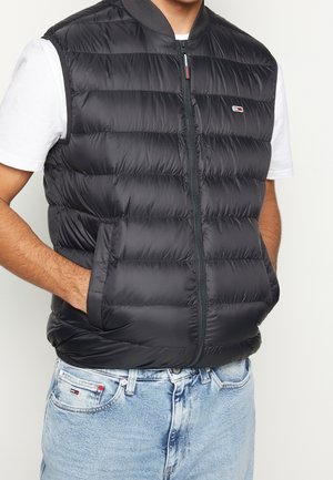 Uomo che indossa un gilet trapuntato nero senza maniche sopra una maglietta bianca e jeans azzurri, con le mani nelle tasche del gilet, in piedi contro uno sfondo bianco.