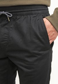 Pantalons de jogger noirs avec une taille élastique, détail à cordon de serrage et poches latérales ; texture lisse avec une coupe ajustée.