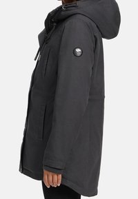 Vue de côté d'une personne portant une veste capuche grise foncée avec fermeture éclair et poches, le poignet côtelé noir visible sous la manche.