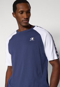 Μπλε t-shirt ναυτικού τύπου με λευκά ραγκλάν μ ανίκια, διαθέτει μικρό λογότυπο στο στήθος και αντίθετες ρίγες με κόκκινες λεπτομέρειες κατά μήκος των μανικιών.