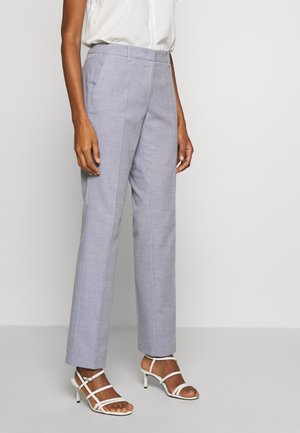 Vrouw met lichtpaarse, op maat gemaakte broek, witte mouwloze blouse en witte sandalen met hak en bandjes, staand tegen een effen achtergrond.