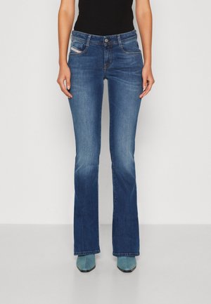Kvinde iført blå bootcut jeans, sort top og turkis spidse sko stående mod en hvid baggrund.