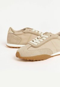 Scarpe sportive beige realizzate in materiali scamosciati e tessili, con lacci bianchi e una suola in gomma texturizzata, esposte su uno sfondo chiaro.