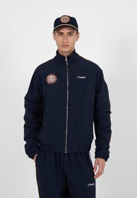 Homme portant une veste zippée bleu marine et un pantalon assorti avec des patchs et le texte « Interdit », portant également une casquette assortie.