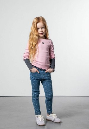 Dziewczyna o długich rudych włosach stoi z rękami w kieszeniach, ubrana w różowo-szary longsleeve, niebieskie jeansy w wzory oraz białe trampki.