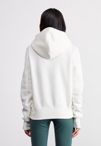 Vit, huva sweatshirt med en mjuk textur; har ribbade ärmmuddar och nederkant. Visad bakifrån, tillsammans med gröna leggings.