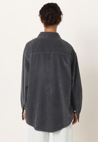 Oseba z zavitimi lasmi, spetimi v nizko figo, nosi preveliko temno sivo kordurovo srajco in svetle hlače, stoji pred enobarvnim ozadjem.