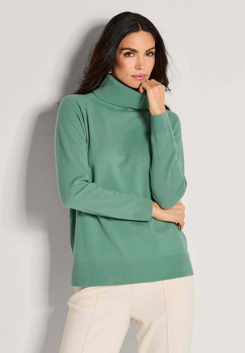 Grüner Rollkragenpullover aus weichem Material, mit einer lockeren Passform, gerippten Bündchen und Saum, kombiniert mit hellen Hosen.