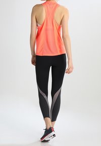 Helles Korallen-Trainingsoberteil mit Racerback-Design, kombiniert mit schwarzen Leggings mit grauen Akzentpaneelen. Schwarze Sportschuhe vervollständigen den Look.