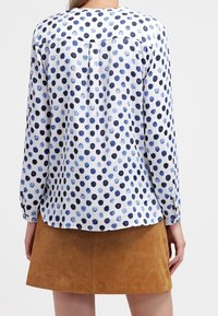 Blusa a maniche lunghe con pois blu e neri su sfondo bianco, caratterizzata da un orlo curvo e un dettaglio con bottoni sul retro. Indossata con una gonna color sabbia.