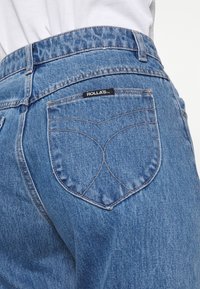 Vue arrière d'un jean en denim bleu avec une étiquette de marque "ROLLA'S" sur la poche arrière droite et des détails de couture visibles.