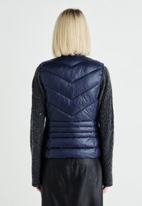 Navy blue puffer vest with chevron quilted design, featuring a lightweight, glossy texture and a high collar. Worn over a grey sweater.