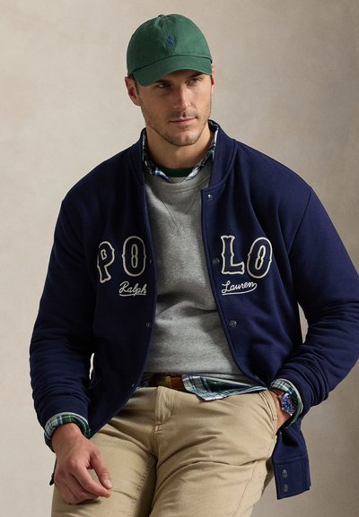 Άνδρας που φορά πράσινο καπέλο, μπλε σκούρο μπουφάν Polo Ralph Lauren πάνω από γκρι φούτερ και πουκάμισο καρό, μπεζ παντελόνι, με το χέρι στην τσέπη.