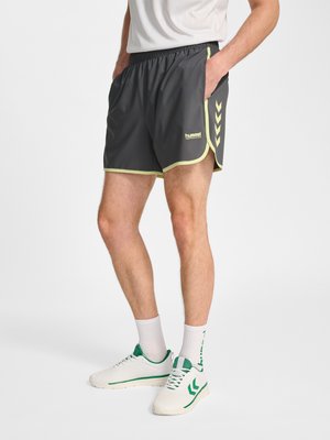 Mężczyzna w czarnych sportowych szortach z żółtym wykończeniem i białych trampkach z zielonymi akcentami, stojący z rękami w kieszeniach.