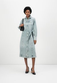 Light blue denim dress with a belted waist, button-front closure, and two chest pockets. Model paired with black heels and a small handbag.