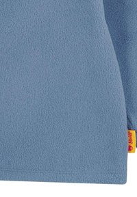 Fleece pulovr v světle modré barvě, s měkkým, texturovaným povrchem a žlutou etiketou s červeným textem na spodním okraji.