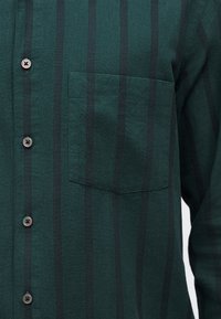 Σκούρο πράσινο πουκάμισο με μαύρες κάθετες ρίγες, διαθέτει τσέπη στο αριστερό στήθος και τέσσερα εμφανή κουμπιά στο μπροστινό μέρος που ταιριάζουν.