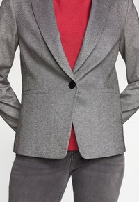 Szary dopasowany żakiet o teksturowanym wykończeniu, z zapięciem na jeden guzik i klapami, nałożony na czerwony top. Widoczny detal z czarnym guzikiem.