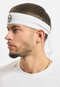 Bílá čelenka s logem, kterou nosí muž. Krátké vlasy, tetování na krku, zlatý řetízek a nosní kroužek, zobrazený v detailním bočním profilu.