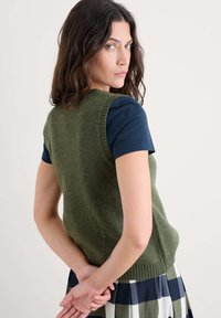 Zaļa vilnas veste ar ribotām malām, kārtota uz tumši zila īsām piedurknēm krekliņa. Nēsāta ar rūtainām svārkiem, kas parāda teksturētu audumu.