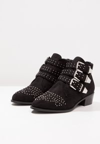 Botines de ante negro con tres correas anchas que presentan hebillas de plata y tachuelas decorativas plateadas en la punta afilada. Tacón bajo de bloque.